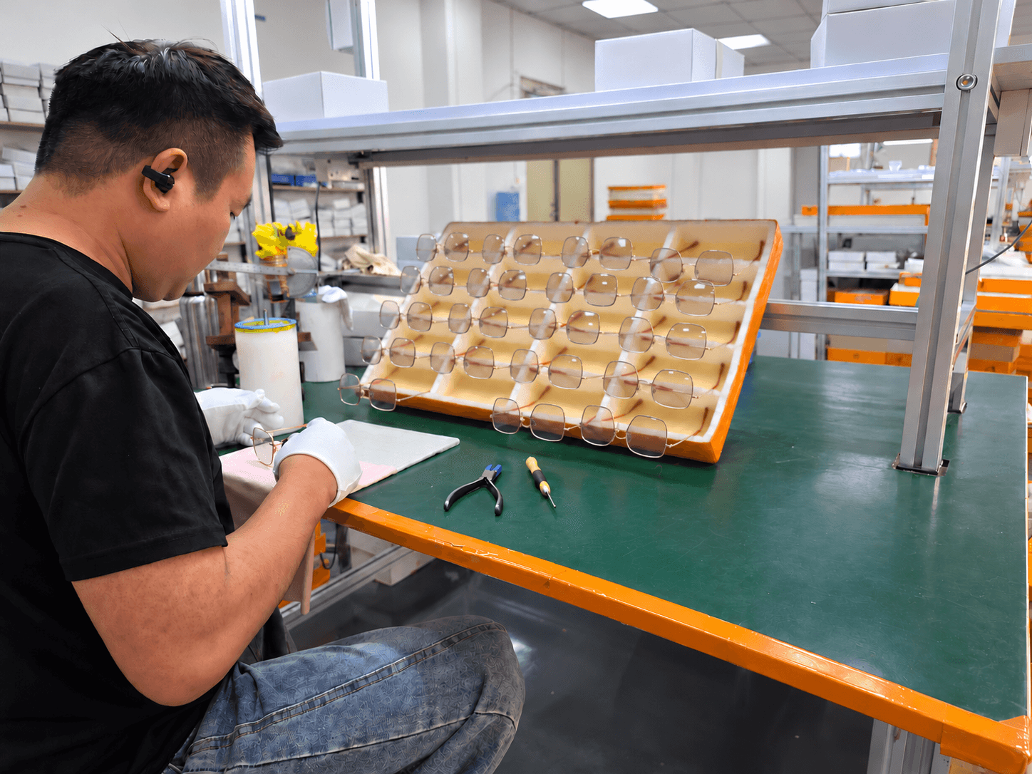 Gloved worker adjusting an eyewear frame at a workshop bench with trays of frames